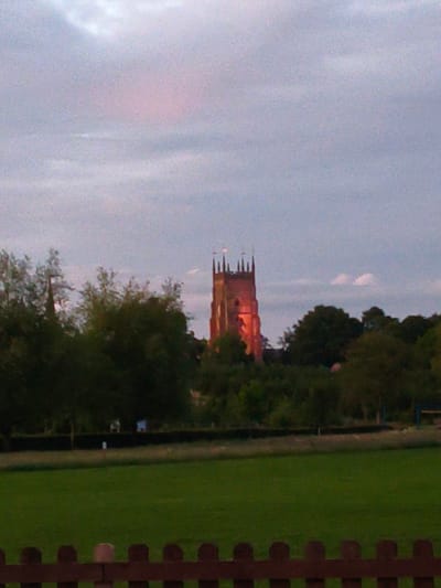 An outdoor, slightly grainy, medium-shot, eye-level, full-color photograph captures a church tower illuminated with a warm, orange glow against a twilight sky. The church, a tall, brick structure with crenellated battlements and pointed spires, is situated in the distance, partially obscured by the dark green foliage of numerous trees that form a dense treeline across the middle ground of the image. The sky above is a soft, hazy mix of light blue and pink hues, with sparse clouds. In the foreground, a dark brown, wooden picket fence runs horizontally across the bottom of the frame. Behind the fence, a lush green lawn stretches into the distance, bordered by a dark hedgerow. The scene appears to be in a park or garden setting. The architecture of the church suggests it could be St. Augustine's Church in Droitwich Spa, within the Wychavon district of Worcestershire, United Kingdom, given its distinctive shape and the likely rural or semi-rural context. No people are visible in the image.