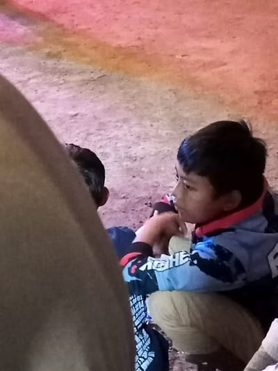 Two young boys are seen sitting on unpaved ground in Pangalengan, Indonesia. The boy on the right is shown in profile, his gaze directed intently towards the right side of the frame. He has dark hair and is dressed in a blue, black, and red hooded jacket. Visible white text on his sleeve includes "ACRE HE", "A#02", and "A#04", with other partially obscured text like "MR." further down the jacket. He is also wearing light brown or khaki-colored pants and appears to be holding a small item or food in his hands.
The second boy is partially visible behind the first, largely obscured by a large, out-of-focus brown object in the immediate foreground, which seems to be a person's leg or arm. Only the back of his head and a portion of his shoulders are discernible.
The ground beneath them is rough and appears to be dirt or gravel. The background is softly blurred with a striking gradient of pink, orange, and light purple light, suggesting an evening or nighttime setting illuminated by colorful artificial light sources. The overall impression is one of quiet observation or engagement, with the visible boy appearing focused on something outside the frame.