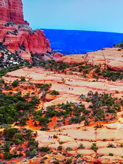 This is an aerial shot of a vibrant desert landscape. A towering red rock formation dominates the left side of the frame, its layered surfaces etched by time and erosion. In the mid-ground and foreground, undulating red rock formations stretch across the scene, dotted with hardy green shrubs and small trees. A clear blue sky forms the backdrop, with a distant blue mountain range visible on the horizon. Several small groups of people can be seen walking on the rock formations, suggesting they are hikers or tourists exploring the area. The overall impression is one of rugged natural beauty and a sense of vastness. There is no information in the image that suggests a location in Gorzow, Poland.