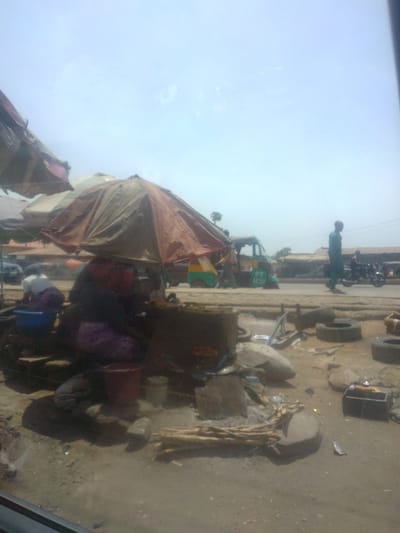 This image depicts a bustling roadside scene in Mista Ali, Nigeria, under a bright, hazy sky. In the foreground, a dirt and gravel surface is littered with various objects, including stacked firewood, metal fragments, and multiple discarded vehicle tires.
Centrally, a person in dark and purple clothing, with a red head covering, is seated beneath a large, weathered brown and orange umbrella, attending to a substantial, dark cylindrical cooking apparatus. To the left of this central figure, another person in white and dark attire is crouched, engaged with a blue basin. Adjacent to the cooking area, a red bucket and a lighter-colored container are visible on the ground, alongside scattered cooking implements.
A paved road occupies the mid-ground, where a green and yellow three-wheeled auto-rickshaw (tuk-tuk) bearing the identifier "PT 1610" is either parked or in motion. A person in light green attire walks from right to left along the roadside, while another individual is partially visible near the tuk-tuk. Further down the road to the right, a motorcycle with a rider is present.
In the background, a row of low-rise buildings lines the far side of the road, suggesting a commercial or residential area. Multiple other market umbrellas are partially visible, contributing to the impression of an active marketplace. The overall environment suggests an outdoor commercial setting with ongoing daily activities.