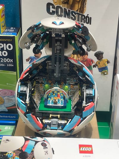 The image depicts a close-up, eye-level shot of a large LEGO soccer stadium model. The model is designed to resemble an open soccer ball, revealing a detailed stadium interior with a green field, bleachers filled with spectators, and a central trophy presentation area. Three LEGO minifigures, two appearing to be soccer players and one referee, are positioned on the exterior of the model. The LEGO set is displayed on a wooden surface with a box of Clementoni educational toys and other retail packaging visible in the background. The overall scene is likely within a retail environment, possibly a toy store or department store, in Rio de Mouro, Portugal.