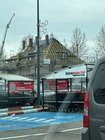 The image displays an outdoor scene, likely a commercial area, on what appears to be an overcast day. The original image is rotated 90 degrees counter-clockwise, making the horizon appear vertical. Describing it as if properly oriented:
On the left side of the frame, a building is undergoing significant construction or renovation. Its roof structure is prominently visible, showing a crisscross pattern of yellowish wooden battens over a blue tarp that covers parts of the roof. Scaffolding surrounds the building, and a white sign attached to the scaffolding reads "Wolff & Chall" and "KARNEV". In the far lower-left background, the arm of a large crane extends upwards against the pale, cloudy sky.
In front of the building, a tall black pole stands, from which a decorative element featuring three illuminated star shapes extends. Bare tree branches with no leaves are scattered across the upper-left portion of the image, suggesting winter or early spring. One tree branch appears to hold a bird's nest.
To the right of the construction site, a supermarket cart return or storage area is visible. This structure holds numerous shopping carts and features several large signs. The topmost sign is a white banner displaying "Intermarché SUPER" in red and black lettering. Below this, a red sign reads "marché d'Ober" and states "MOINS DE PAPIER, ET TOUJOURS AUTANT DE PROMOS." (less paper, and always as many promotions) along with a QR code. Another similar red sign is partially visible below it, also containing text and a QR code.
On the far right of the image, the rear passenger side of a grey van or car is visible. The word "PARTNER" is printed in white letters on its side, and a red taillight is clearly seen. The ground in the immediate foreground, to the right, is part of a parking area. A section of the ground is painted bright blue with white markings, possibly indicating a designated parking spot. An orange and white striped curb or barrier separates this blue area from other ground surfaces.