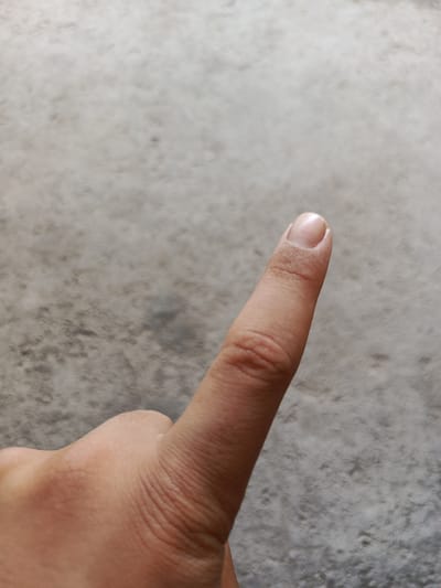 A close-up, overhead view of a person's index finger and thumb is visible against a textured, greyish-brown surface, likely concrete or stone. The finger is pointing upwards and slightly to the right. The skin on the finger shows faint wrinkles and has a light tone. The fingernail is visible at the tip of the finger, appearing clean and smooth. The thumb is partially visible in the lower left corner of the frame. The background is out of focus and uniform in color and texture. There is no indication of the city of Guba, Azerbaijan, or any specific urban environment.