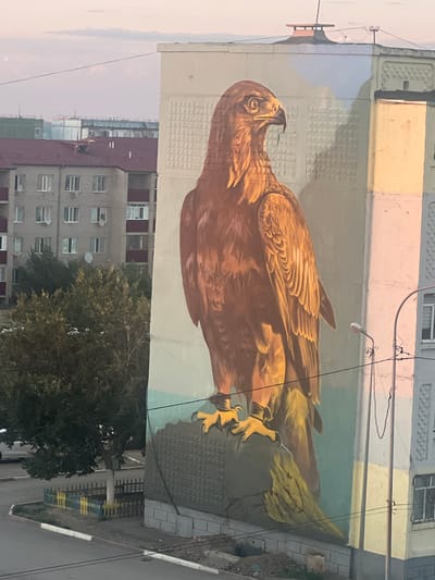 A mural of a brown bird, possibly a falcon or eagle, is painted on the side of a building. The bird is depicted in profile, facing left, with its wings partially spread and its talons extended. The mural uses warm earthy tones for the bird, with contrasting blue and green colors in the background, possibly representing sky and foliage.
The building itself appears to be an apartment complex, with visible windows and balconies. In the foreground and background, other buildings are visible, some with red and grey facades. Trees with green foliage are present in front of some of the buildings. Electrical wires and poles are also visible, suggesting an urban or suburban environment. The lighting suggests it might be early morning or late afternoon, with a soft, diffused light.
Below the mural, a street with a grey asphalt surface is visible, lined with a low fence made of yellow and blue elements. There are no people clearly visible in the image, and the overall mood is serene, focusing on the art and the surrounding architecture. The location is identified as Satbayev, Kazakhstan. No visible text is present in the image.