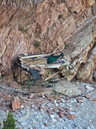 A vertical, eye-level, outdoor shot captures a crude, unfinished wooden structure constructed against a sheer, reddish-brown rock face at Perazića Do, Montenegro. The structure is supported by green metal poles and features a stacked pile of planks, some weathered and others showing signs of colorful paint. A dark green tarp is draped over part of the construction, and debris, including more wood, rocks, and what appears to be wire mesh, litters the ground around it. The rock face is characterized by distinct horizontal striations and pockets of greenery. The ground in the foreground is a mix of pebbles, larger rocks, and sand. A thin, white cable runs vertically along the rock face to the left of the structure. The overall scene suggests a work in progress or a derelict site.