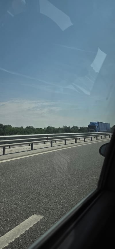 The image depicts a daytime scene viewed from the interior of a moving vehicle, likely a car, looking forward and slightly to the left. The setting is a multi-lane highway or motorway in Trilistnik, Bulgaria.
The upper portion of the frame shows a pale blue sky with scattered light clouds and several faint contrails. Prominent reflections, likely from the car's interior or external elements, are visible across the upper right portion of the window, obscuring parts of the sky.
The road surface in the foreground and midground is dark grey asphalt, textured, with a solid white line defining the right edge of the visible lane. To the left of this line is a lighter, unpaved or gravel shoulder/median area. A silver, double-bar metal guardrail runs parallel to the road on the left, supported by vertical posts.
Beyond the guardrail, a dense line of green trees extends across the horizon, indicating a rural or vegetated environment.
A blue semi-trailer truck (lorry), identifiable as a modern Scania model, is visible in the distance on the left side of the road, traveling in the same direction or on an opposing lane of a divided highway.
The right edge of the frame shows the dark interior molding of the car's window and possibly the housing of a side-view mirror. The overall lighting suggests bright daylight conditions.
