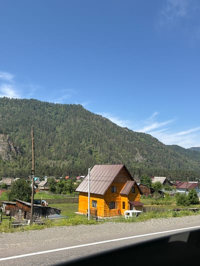 The image depicts a bright, sunny day in the village of Cheposh, Russia, characterized by a clear blue sky with wispy clouds. A prominent feature is a vibrantly orange, two-story house with a brown, pitched roof, situated in the foreground. To the left of the house are several older wooden structures, some appearing weathered and in disrepair, alongside a tall wooden utility pole with visible wires.
The village is nestled against the base of a densely forested mountain that dominates the background. The mountain's slopes are covered in a thick canopy of evergreen trees. In the mid-ground, more houses with various roof colors and styles are visible, interspersed with green grassy areas and wooden fences. A paved road with a white dividing line runs along the very bottom of the image, suggesting the perspective is from a vehicle or roadside. The overall environment is rural, with a strong sense of natural landscape integrated with a small settlement.