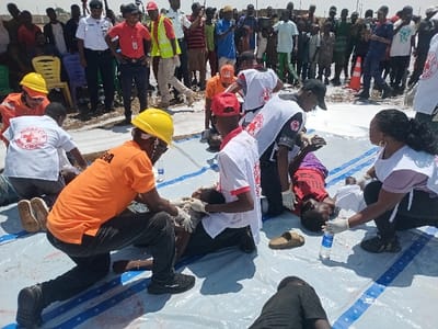 The scene is an open area in Katabu, Nigeria, near latitude 10.71 and longitude 7.514. It is a medical aid station with several people working to help an injured person. A man wearing a yellow hard hat and an orange shirt kneels and examines the patient. A woman with long, dark hair and a white shirt kneels on the opposite side of the patient while holding a white cloth. Another man with a red cross shirt and a red hat sits next to the patient's head, while two other men with red cross shirts and red hats kneel near the patient's legs. The patient is lying on their back, wearing a purple shirt, with their left hand lying on the ground and their right hand raised. A second man with a red cross shirt and a red hat watches the scene, standing near the patient. A large group of people stand and watch the scene from a distance, most wearing brightly colored clothing. There are several objects in the scene, including plastic chairs, water bottles, a white tarpaulin sheet, and an orange traffic cone. The surface of the ground is a white, dusty, open area. The scene is well lit by the bright sun shining on the open area.
