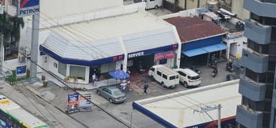 The image shows a street view of a Petron gasoline station with a service center in Makati, Philippines. A prominent PETRON sign is visible. The service center has a white roof and blue trim. Several vehicles are present, including a white van with a roof rack, a white SUV, and a silver sedan. A bus is partially visible on the left side of the image. People are seen walking and interacting in various areas, including near the service center and on the sidewalk. A blue canopy tent is set up in front of the service center. Price boards displaying fuel prices are positioned near the road. The environment appears to be an urban setting with buildings and overhead wires.