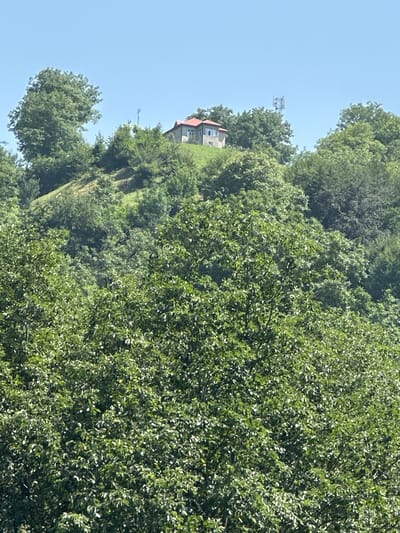 The image shows a hilltop with a house and a communication tower, surrounded by lush green trees. The house has a red roof and is situated on a gentle slope. The communication tower, a tall metal structure, is visible in the background. The sky above is clear and blue. The environment suggests a rural or semi-rural setting, potentially in the city of Kusnatqazma, Azerbaijan, given the context. The foreground is dominated by dense foliage, obscuring the lower parts of the scene. There are no people visible in the image. The overall impression is one of a tranquil landscape with human structures integrated into the natural environment.