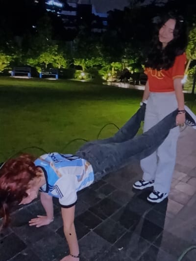 The scene depicts two individuals at night on a dark paved surface adjacent to a grassy, illuminated park area in Baykaro, Uzbekistan. The individual in the foreground, with reddish-brown hair and wearing a light blue and white striped "MESSI 10" jersey and dark jeans, is in a plank-like position, supported by her hands on the pavement. Her legs are held by a second individual standing behind her. This second individual, with long dark hair, is wearing a red short-sleeved top with a yellow design, light-colored pants, and black and white sneakers. She holds the first individual's lower legs at the ankles. The paved surface consists of dark, rectangular tiles. In the background, a manicured grassy area is visible, brightly lit by ground-level lights, illuminating trees and bushes. Two blue park benches are discernible within the grassy area. Distant, dark building silhouettes are visible against the night sky above the treeline. The interaction involves a physical exercise or playful pose, with one person supporting the other in a "wheelbarrow" position.