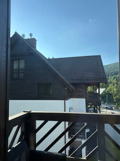 The image shows a view from a wooden balcony overlooking a building with a dark, tiled roof and dark wooden siding. The building has several windows and what appear to be ventilation pipes on the roof. To the right of the building, there is a portion of the balcony with diagonal wooden supports. Beyond the building, a road is visible with cars driving on it, and a forested hillside rises in the background under a clear blue sky. The sun is bright, casting sharp shadows, indicating it is likely midday. The scene appears to be a peaceful, scenic location in Szklarska Poręba, Poland. There are no people visible in the image.