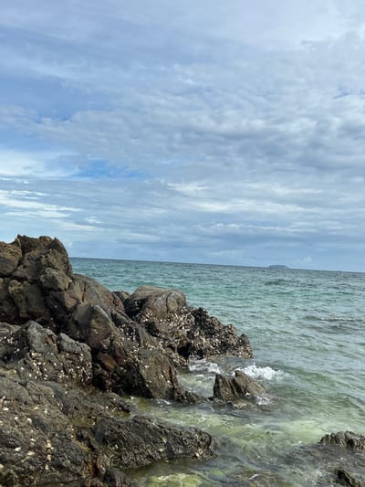 This media file captures a scenic coastal view in Tambon Phru Nai, Thailand. The image is oriented vertically, presenting a dynamic split between the sky and the sea. The left side of the frame is dominated by a dramatic sky filled with layered clouds. These clouds range from soft, wispy formations to thicker, more textured bands, suggesting a day with variable weather. The hues of the sky are predominantly blues and whites, with hints of grey indicating the cloud cover.
The right side of the image depicts the ocean and a rocky shoreline. The water is a pale, translucent green, with gentle waves lapping against dark, rugged rocks. These rocks are covered in barnacles and other marine life, adding a textured and natural element to the scene. The water appears calm, with small whitecaps indicating the movement of the waves. In the distance, a faint outline of land or an island can be seen on the horizon, partially obscured by the haze.
The overall mood is one of natural beauty and tranquility. The light suggests it could be daytime, possibly late morning or early afternoon, given the brightness but also the cloud cover. There are no people or man-made objects explicitly visible, emphasizing the raw, natural environment. The scene conveys a sense of peacefulness and the vastness of nature. No text is visible in the image.