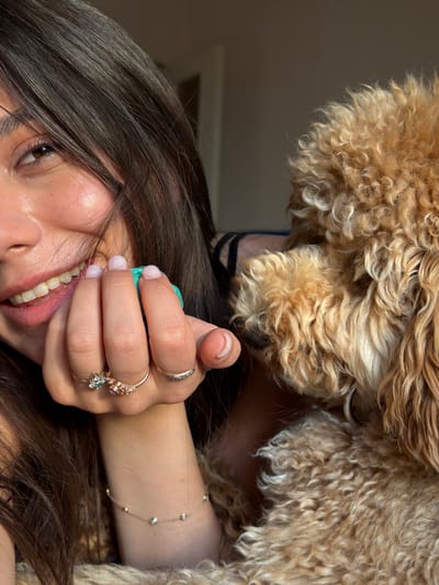 The image shows a close-up of a woman with dark hair and a light-skinned complexion, smiling and holding a turquoise object near her face. She is wearing several rings on her fingers, including one with green and purple stones and another with a wave-like design. A delicate gold anklet with small beads is visible on her arm. Beside her is a fluffy, light brown dog. The lighting suggests an indoor setting, possibly with natural light coming from the side. There is no indication of the location being in Aratashen, Armenia.