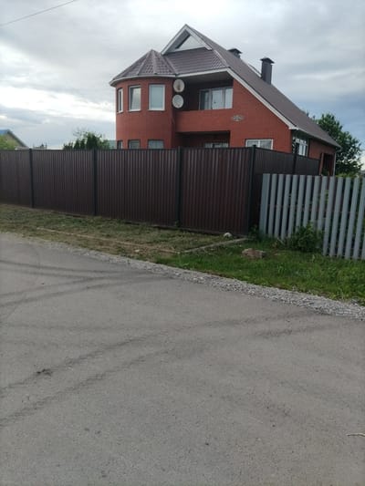 This image depicts a residential street scene in Nizhnyaya Makhtama, Russia, under an overcast sky. The main subject is a two-story brick house with a dark brown metal fence running along the front. The house has a complex roofline with dormer windows and a prominent chimney. Two satellite dishes are visible on the exterior of the house. The fence is made of dark brown corrugated metal panels, with a section of lighter gray metal vertical panels to the right.
The foreground of the image shows a paved asphalt road with tire marks, indicating recent vehicle activity. The road is bordered by a gravel strip and a patch of grass and weeds. The setting appears to be a suburban or rural area, with other houses and trees visible in the background beyond the fence. The weather appears to be cloudy and possibly humid, given the subdued lighting. There is no discernible activity or people in the frame. A small sign with Cyrillic text, possibly a house number or street name, is affixed to the brick wall of the house above the fence.