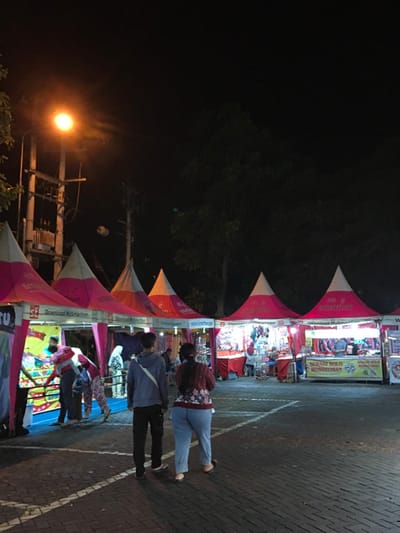 The image shows a night market in Kecamatan Batu, Indonesia, located at latitude -7.867 and longitude 112.514. The scene is illuminated by streetlights. There are five tents lined up in a row, each with a white top and pink sides. The tents are selling a variety of goods. There are several people walking around and browsing the stalls. Some people are shopping and others are looking at the goods. There are two men and two women. One of the women is wearing a long-sleeved shirt and pants. The other woman is wearing a black long-sleeved shirt and dark jeans. One of the men is wearing a blue shirt and jeans, while the other is wearing a gray shirt and pants. The ground is covered in brick pavers.