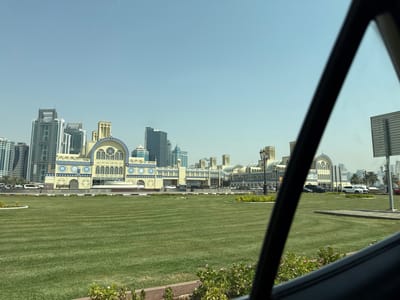 The image shows a view of the Blue Souk, also known as the Sharjah Old Souk, located in Sharjah, United Arab Emirates. The Blue Souk is a prominent landmark with its distinctive blue-domed architecture. In the foreground, a meticulously maintained lawn with striped patterns stretches out. Behind the lawn, the Blue Souk's facade is visible, characterized by its arched windows, intricate blue and yellow decorative motifs, and minaret-like towers. The structure is flanked by modern skyscrapers in the background, indicating the coexistence of traditional and contemporary architecture in the city. A clear blue sky forms the backdrop. The perspective suggests the photo was taken from a vehicle, with a portion of the car's interior, including a dark-colored frame, visible in the foreground. Several cars are parked and driving on the road in front of the souk.