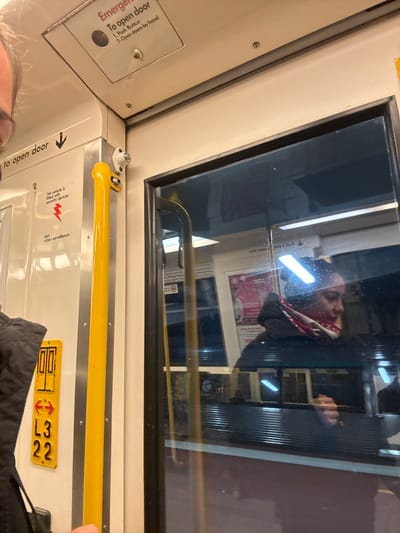 This appears to be an interior shot of a train car, specifically near a door. The emergency door release mechanism is visible, with instructions indicating a push button and manual operation. There is a yellow grab bar, and a sign indicating that the vehicle is fitted with security devices and video surveillance. A yellow sign displays "L3 22" and an arrow, possibly indicating the train car's location or identification. A reflection in the window shows an obscured image of a person, and the interior of the opposite side of the train. The precise location in Bassendean, Australia, cannot be determined from the image alone.