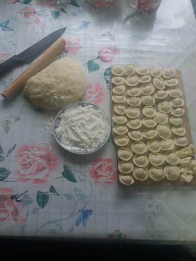 The image depicts a food preparation scene, likely indoors. In the upper portion of the frame is a wooden cutting board on which approximately 50 pelmeni (small dumplings) are arranged closely together. Below the board, a small ceramic bowl containing a mound of white flour sits on the table. To the left of the bowl, a ball of dough rests next to a black-bladed knife and a wooden rolling pin. The surface beneath these items is a tablecloth or covering with a floral pattern of pink roses and butterfly motifs. The location is most likely a kitchen or food preparation area.
