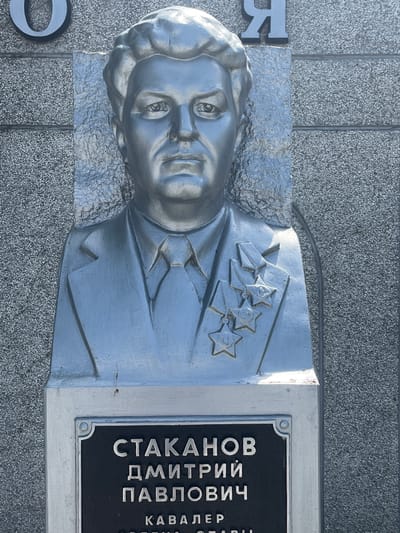 This media file features a close-up, vertical shot of a silver bust of a man, likely a monument. The bust is positioned on a gray, textured surface that resembles granite, with vertical lines suggesting it's part of a larger structure or wall. Behind the bust, the background is also a gray, speckled texture, possibly the same granite material.

The bust depicts a man with short hair, wearing a uniform adorned with several star-shaped medals on his chest. He has a stern expression, with his eyes looking forward. The detail on the face, hair, and clothing is intricate, with shadows and highlights adding depth to the metallic finish.

To the right of the bust, a black plaque with engraved silver text is visible. The text is in Cyrillic script and appears to be a name and possibly a title or rank. Partially visible text reads: "СТАКАНОВ ДМИТРИЙ ПАВЛОВИЧ" and below it, "КАВАЛЕР". The phrase "КАВАЛЕР" suggests he was a knight or a bearer of an award.

On the left side of the image, a portion of a large, stylized silver letter "R" is visible, hinting at a potential inscription or monument title. Another partial letter, possibly an "O", is visible at the bottom left corner.

The lighting suggests it is daytime, possibly under direct sunlight, as evidenced by the strong highlights and shadows on the bust. There are no overt indications of weather conditions. The overall scene suggests a memorial or historical site, given the nature of the bust and the plaque. The setting is likely an outdoor public space, possibly a park, square, or cemetery in Takhtamukai, Russia.