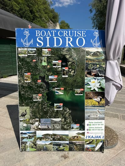 This is a full outdoor shot of a large blue sign advertising boat cruises on Skadar Lake, near Rijeka Crnojevića, Montenegro. The sign features an aerial map of the lake with various locations marked with prices, and smaller inset photos showcasing wildlife and scenery. The sign is propped up on a stone surface, with a grey umbrella visible to the right. The background shows lush green foliage and a bright blue sky with some clouds. There are no people visible in the frame.