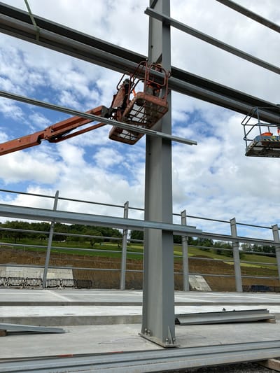 A construction site in Wilkowisko, Poland features the skeletal framework of a building under assembly. Two cherry pickers, one orange and one blue, are positioned near large steel beams. The orange cherry picker is being operated by a construction worker wearing a hard hat and safety vest. The worker in the blue cherry picker is obscured. The beams are suspended in the air, supported by a large vertical steel column in the foreground. In the background, there is a grassy slope with trees, and a concrete foundation. The sky is cloudy with patches of blue.