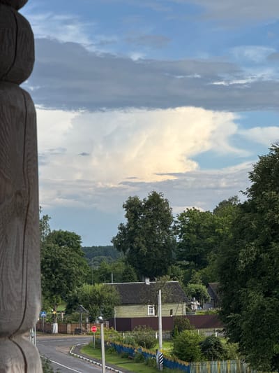 The image depicts a rural landscape under a cloudy sky, likely in the vicinity of Нёманскі сельскі Савет, Belarus. The foreground on the left features a portion of a weathered wooden structure, possibly a porch or balcony railing, with visible grain and texture. The mid-ground reveals a village scene. A light green, weathered wooden house with a dark roof and a small white-paned window is visible. Adjacent to it, a dark brown fence encloses a property. A paved road curves through the scene, marked by white lines and bordered by a black and white striped curb. A red octagonal STOP sign stands on a pole near the road, accompanied by a blue and white pedestrian crossing sign. Colorful painted wooden fences, with alternating blue and yellow pickets, line the roadside. Various trees, predominantly deciduous, are scattered throughout the mid-ground and background, with a denser forest covering the hills in the distance. The sky is dominated by dramatic clouds, with a large, bright, illuminated cloud formation in the center, suggesting a break in the weather or the approach of a storm. The overall impression is one of a quiet, overcast day in a rural Belarusian settlement.