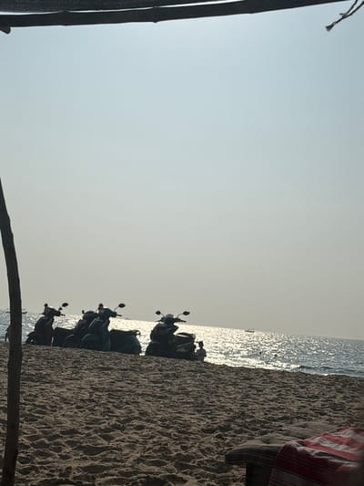 The image displays a beach scene in Bhagatwadi, India, under bright, possibly late afternoon, natural light. The foreground consists of textured sand, leading towards the ocean. On the lower right, a striped fabric, primarily red and white, covers a low, dark-colored object, possibly a cushion or mat. Along the left margin, a dark, vertical support structure, appearing to be a tree trunk or pole, extends upwards. A dark, horizontal beam is visible at the top edge of the frame.
In the midground, five two-wheeled motor scooters are parked sequentially on the sand, silhouetted against the brighter background. Their specific colors are obscured by backlighting, but shapes are discernible. To the right of the rightmost scooter, a small human figure, possibly a child, is seated or squatting on the sand near the water's edge.
The background features a vast expanse of ocean, reflecting strong sunlight that creates a shimmering, bright surface. Several small, dark shapes, consistent with distant boats, are visible on the water, particularly towards the left horizon. The sky above the ocean is clear, hazy, and pale blue-grey, occupying approximately the upper half of the image.