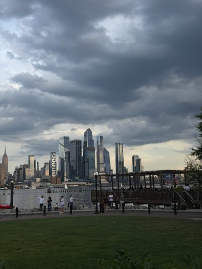 The image depicts a cloudy day in Weehawken, USA, with a panoramic view of the New York City skyline across the Hudson River. In the foreground, a grassy park area is visible, with a pathway bordering a rocky shoreline. Several people are present along the shoreline and on a wooden pier. Some are standing and looking out at the water or the skyline, while others are gathered on the pier, possibly at an outdoor seating area. A sailboat is visible in the water. The skyline features numerous modern skyscrapers, including the Empire State Building on the far left. The sky is filled with dark, dramatic clouds, suggesting an approaching storm or overcast weather.