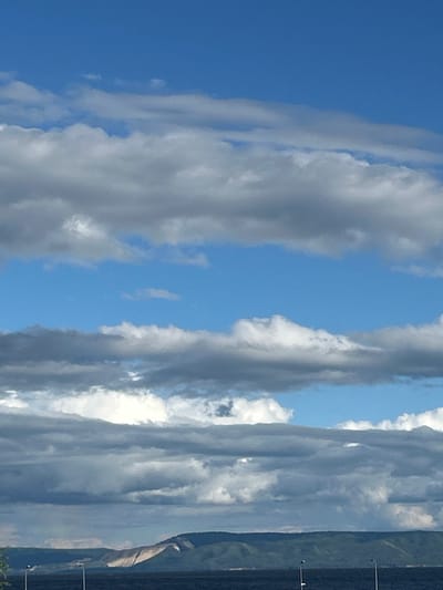 This is a vertical, eye-level, outdoor, daytime photograph capturing a wide expanse of sky over a body of water and distant hills. The sky is a vibrant blue with numerous scattered clouds of varying density and color, ranging from bright white to dark gray. Some clouds are layered horizontally, creating a sense of depth.
In the lower portion of the image, a dark blue body of water stretches across the frame, meeting a distant, rolling landscape of green hills. The hills appear to be covered in vegetation and have a layered, undulating topography. The horizon line is low in the frame, emphasizing the dominance of the sky.
Two minimalist streetlights with thin poles stand in the foreground, their tops cut off by the bottom edge of the image. The light fixtures themselves are simple and dark. To the far left, a hint of dark green foliage is visible at the edge of the frame.
The scene is serene and natural, with no visible people or animals. The location appears to be a waterfront area, potentially a park or promenade, in or near the city of Primorsky, Russia, given the characteristic landscape and cloud formations often seen in the region. The overall impression is one of natural beauty and expansive sky.