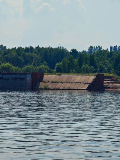 The image depicts a waterfront scene in Ust-Kem, Russia. In the foreground, a body of water with rippling surfaces fills the bottom half of the frame. The middle ground features a concrete structure that appears to be a dam or embankment, with a sloped surface covered in concrete slabs and patches of grass. To the left of this structure, a more industrial-looking concrete wall is visible, with some dark, irregular shapes that could be mooring posts or part of ongoing construction. The right side of the concrete structure transitions into a sandy bank with steps leading up. The background is dominated by a dense line of lush green trees, forming a forest that stretches across the horizon. Above the trees, a light blue sky with scattered white clouds is visible. Towards the right side of the tree line, a tall, slender industrial tower or structure can be seen.