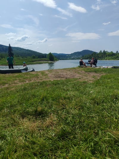 In Mezhdurechensk, Russia, on a clear day with scattered clouds, a group of people are gathered by a wide body of water, likely a reservoir or large river. In the foreground, tall, green grass covers the landscape, with some patches of drier, brown grass. A dirt path leads towards the water's edge.
To the left of the image, a green inflatable raft is partially beached on the shore. A man in a green t-shirt and dark shorts stands near the raft in the shallow water, facing away from the camera. A young child wearing a blue hat stands beside him in the water.
Further along the shore, to the right, three adults are seated in camping chairs on a sandy area. One individual, facing the water, is partially obscured by a table in front of them. Another person, with blonde hair, is seated and looking towards the water. A third person is also seated, with their back to the camera. Several items, including what appears to be a blanket and a bag, are laid out on the ground near the seated individuals.
In the distance, across the water, a tree-covered hillside rises. Hills with scattered buildings are visible further in the background, suggesting the presence of a settlement or town. The sky is a bright blue with fluffy white clouds. The overall scene depicts a recreational day by the water.
