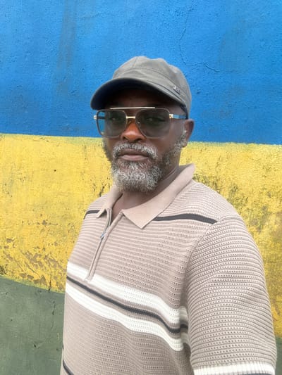 The image shows a close-up of a Black man wearing a grey cap and sunglasses. He has a beard and is looking slightly to the side. He is wearing a light-colored, textured sweater with dark and white stripes on the sleeves and collar. The background is a painted wall with a horizontal stripe of bright blue and yellow. The painting appears to be on an exterior wall. The man is centered in the frame, and the lighting suggests it is daytime, likely sunny given the shadows. The overall mood is casual and relaxed. No visible text is present in the image. The location context provided is Ikoyi, Nigeria.