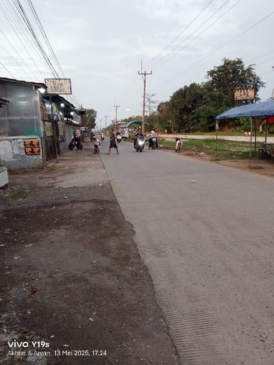 The image shows a street in Kecamatan Kroya, Indonesia, with a gray, paved road in the center. The road is divided by a line of dark soil on the left. There are five motorbikes driving on the road. Three people are walking on the left side of the road. There is a blue awning on the right side of the road. The street is lined with trees and a few buildings on the left side, with a sign reading "TOKO REVAN KOMURELA CELANA". There is a white pickup truck with a blue awning in the distance. The sky is overcast and gray.