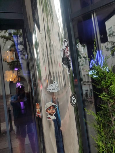 The image shows a reflective metal pole on the exterior of a building, likely a shop or studio, in Rosenheim, Germany. The pole is adorned with several stickers. Among them is a sticker with the text "MOUNT INK tattoo studio" and an illustration of mountains. Another sticker features a cartoonish character with a baseball cap and large blue eyes, and a third sticker depicts a person with black hair and a red outfit. Reflections in the pole reveal the street scene, including buildings and foliage. A neon sign, partially obscured by a green plant, is visible in the background. The overall impression is that of an urban street environment with commercial establishments.