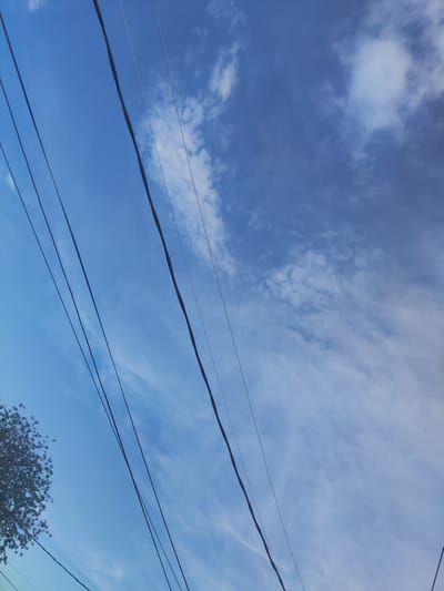The image is a photograph taken outdoors, looking upwards at a blue sky with scattered white clouds. Several power lines are visible, running diagonally across the frame. In the lower left corner, the top of a green tree with branches and leaves can be seen. The scene appears to be an urban or suburban environment, possibly in Manole, Bulgaria, given the context. There are no people or animals visible in the image.