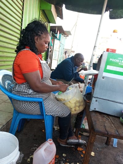 This image, captured in Tala ward, Kenya, depicts an outdoor informal food preparation and sales area. In the foreground, an adult female with dark complexion and dark hair styled in braids is seated on a blue plastic chair, facing left. She wears an orange short-sleeved top and a black-and-white checkered apron over dark trousers. Her hands are engaged in peeling or preparing potatoes from a transparent plastic bucket, which contains numerous cut potato pieces. Organic debris, including potato peels, is scattered on the dark ground beneath her feet. A white plastic bucket is on the ground to her left, and a translucent orange plastic bottle is to her right.

In the mid-ground, an adult male with dark complexion and a bald head is seated on a blue plastic stool, facing right. He wears a dark blue long-sleeved shirt and is focused on a small object or container in his hands, appearing to consume food. To his immediate right, a gray metal vendor cart with a wooden top is visible. A green and white sign affixed to the cart partially reads "LIPA NA POCHI LA..." with a "DIAL..." prompt, indicating a mobile payment service. A yellow plastic bottle and a smaller red bottle are present on the cart's surface. Overhead, a white pole supports a green tarp providing shade. The background features a closed green roll-down shutter of a building on the left, with additional indistinct buildings and a distant human figure further along what appears to be a street or pathway.