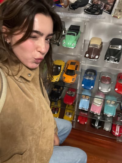 A person wearing a tan suede jacket and blue jeans poses in front of a glass display case filled with toy cars. The collection includes various models, colors, and styles. The person's face is close to the camera, with lips pursed as if giving a kiss. The focus is split between the individual and the car collection behind them. The background includes a wooden surface to the right of the display.