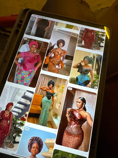 A tablet screen is the central focus, displaying a grid of nine smaller images, predominantly featuring women in elaborate formal attire. The tablet, possibly situated in Dushepe, Nigeria, is dark-cased and held at an angle, with its screen illuminated against a dimly lit background.
The displayed images showcase women adorned in richly colored and embellished dresses, characteristic of Nigerian formal fashion. Outfits include a deep red gown with intricate details, a teal and black dress with large bows, a shimmering copper-toned dress, an orange and gold ensemble, a pink lace top paired with a vibrant Ankara skirt, and a mint green sequined gown. Many women wear traditional headwraps (gele) in coordinating colors, and some accessorize with jewelry like earrings and bangles. The women are often posed elegantly, suggesting a fashion showcase, portfolio review, or social media browsing activity.
Visible text on the tablet includes a timestamp "19:09 Wed. 29 Apr" indicating the tablet's system time. Individual images also bear timestamps such as "0:45," "0:35," and "0:16," suggesting these may be video clips. The caption "Lemon Made" is also partially visible on one image. The overall scene highlights a collection of stylish, culturally rich fashion designs, possibly from a designer or an event.
