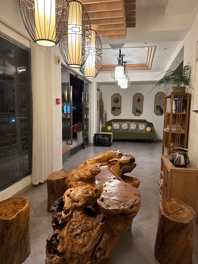 The image depicts an interior space in Zhuoshui, China, featuring a unique aesthetic combining natural wood elements and contemporary design. A large, gnarled wooden structure dominates the foreground, likely serving as a table or decorative element, surrounded by cylindrical wooden stools. A green sofa adorned with patterned pillows sits against a wall decorated with framed botanical prints. Overhead, hanging lamps with woven shades cast a warm light. The background reveals a glass-enclosed area with floral arrangements and a glimpse of outdoor scenery. Shelves stocked with books and decorative items are visible, suggesting a curated, living space. The flooring is a light-colored tile, complementing the wood tones.
