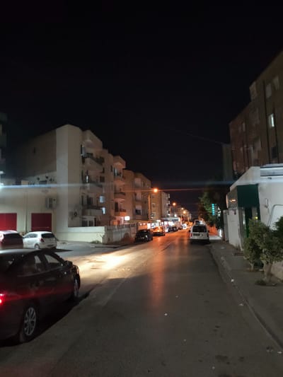 The image depicts a nighttime street scene in Unknown, Tunisia. The perspective is from a street level, looking down a road with buildings lining both sides. The sky is dark, indicating it is night.
On the left side of the street, there are multi-story apartment buildings with visible windows, some illuminated. There are several parked cars, including a dark-colored sedan in the foreground on the left, and a white hatchback and another car parked further back. A building with large red doors is also visible on the left.
On the right side of the street, there are more buildings, including a building with a white facade and a dark green structure. Vegetation, such as bushes and a small tree, is present along the sidewalk. There are streetlights casting illumination on the road.
Several vehicles are on the road, including a white van with visible license plates, and other cars further down the street, some of which have their headlights on. The road surface appears to be asphalt, with some debris visible. The overall atmosphere is that of a quiet urban street at night.