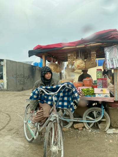 This media file captures an outdoor scene, likely a street or market stall, in Nakhonay, Afghanistan. The central subject is a young person, possibly a boy, sitting on a silver bicycle. He wears a dark jacket, brown trousers, and a black and white checkered head covering. A blue and white checkered blanket is draped over the handlebars of his bicycle. He is looking towards the viewer with a neutral, slightly pensive expression.

To the left of the person is a street vendor's cart or stall, constructed with a wooden frame and covered by a red and black tarp acting as an awning. The cart displays a variety of goods, including packaged snacks like chips or flatbreads in clear plastic bags, containers of colorful sweets or candies, and possibly dried fruits or nuts in clear plastic jars and boxes. Another person, partially obscured, is visible behind the vendor's cart, wearing dark clothing.

The setting appears somewhat rustic, with a dirt or muddy ground. In the background, there is a plain, rough-textured concrete or mud wall. Visible on this wall is script in Farsi or Dari, which includes "هومستای پلان" (possibly "Homestay Plan" or a similar phrase) and a phone number: "0202051572". Behind the wall, two blue plastic barrels or containers are faintly visible. The sky is overcast and bright grey, suggesting daytime but with no direct sunlight. On the far right, part of a rustic, possibly broken, cart or trolley with wheels is seen on the ground.