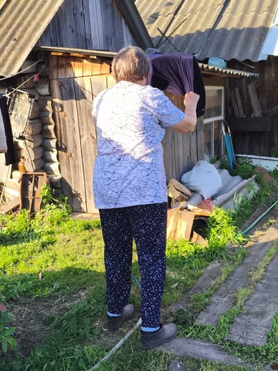 A woman is standing in a yard in front of a wooden structure in Ilmino, Russia. She is wearing a white t-shirt with a black abstract pattern and dark blue pants with a white star pattern. She has on dark rubber boots. She is holding a dark blue garment, possibly clothing, up in her hands. The wooden structure appears to be a shed or a small building with a corrugated roof. There is a pile of white bags and wood scraps near the structure, and the yard is covered in green grass and some dirt paths. The sun is casting shadows on the scene, indicating it is daytime.