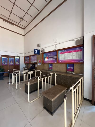 The image shows the interior of a ticket counter area at a train station in Desa Tambakreja, Indonesia. The scene is viewed from a low angle, looking towards the counter. The walls are white, with brown trim along the ceiling and corners. A grid-like ceiling structure with brown lines is visible. Fluorescent lighting is present in the ceiling.
In the center of the image, a long counter with a textured brown surface is visible. Behind the counter, there are several digital displays showing train schedules and information. One prominent display reads "JADWAL KERETA API" (Train Schedule). The displays are framed in wood. Several potted plants are located to the left of the counter, near wooden double doors.
In front of the counter, there are metal railings forming a queueing system. These railings are painted white and show signs of wear, with chipped paint in some areas. The floor is tiled with a light-colored material, and grid lines are visible, indicating the grout lines between the tiles.
One individual, dressed in a dark blue shirt, is visible behind the counter, appearing to be serving a customer or attending to the displays. The person's face is not clearly visible. A sign with a blue icon and text, possibly indicating a ticket counter, is mounted on the wall above the counter. Another sign with the number "121" is also visible near the counter.
Posters with images of luggage and text are displayed on the wall above the counter, likely providing travel information or advertisements. The overall impression is that of an operational train station ticket counter.