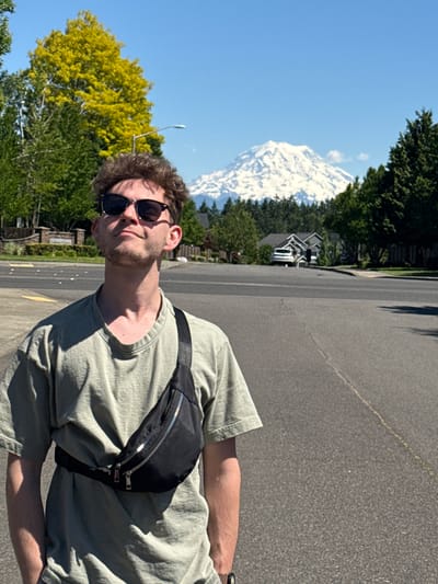A young man with curly brown hair and sunglasses stands in the middle of a paved road in South Hill, USA, with a snow-capped mountain in the background. He is wearing a light green t-shirt and a black fanny pack across his chest. The sun is shining brightly, casting shadows on the road and the man. Trees and houses are visible on either side of the road. The mountain is large and imposing, with snow covering its peak and slopes. The sky is a clear blue.
