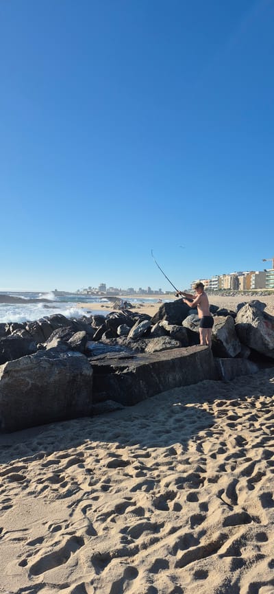 This vertical image depicts a sunny day at the beach in Vila do Conde, Portugal, with the Atlantic Ocean in the background. A young, shirtless male is standing on a rocky jetty, actively fishing with a long rod. He is wearing dark swimming trunks and a light-colored cap. The ocean exhibits choppy waves crashing against the rocks, with a clear blue sky overhead. In the distance, the cityscape of Vila do Conde is visible, featuring buildings and a lighthouse. The foreground is dominated by a sandy beach with footprints. The scene captures a moment of recreational activity against a coastal urban backdrop.