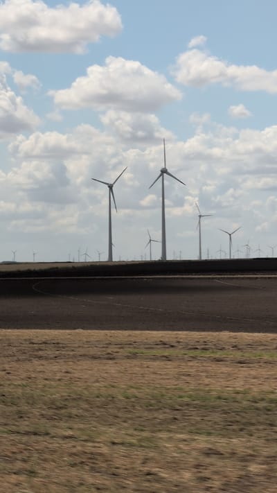 The image depicts a landscape with multiple wind turbines, a cultivated field, and a cloudy sky. The wind turbines, which appear to be the primary focus of the scene, are positioned in the background, scattered across a horizon line. They are white and tall, with their blades extended, suggesting they are operational. In the foreground, there are two distinct fields. The field closer to the viewer is characterized by dry, straw-colored vegetation with hints of green, and it is noticeably blurred, indicating motion. Behind this, separated by a sharp line, is a darker, plowed field with visible furrows. The sky is partly cloudy, with white cumulus clouds against a pale blue background. There is no discernible urban infrastructure or any indication of the city of Baia, Romania, in the image. The scene is rural, focusing on agricultural land and renewable energy infrastructure.