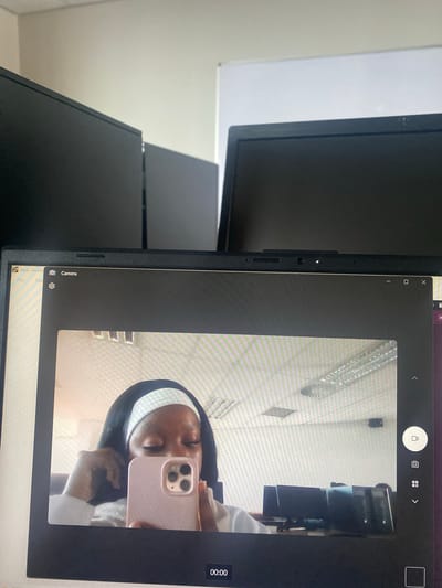 The image shows a laptop screen displaying a camera application with a selfie of a person holding a pink phone. The person in the selfie has dark hair with a white headband. The background in the selfie is a white wall with a ceiling fan. The laptop is surrounded by multiple computer monitors. The environment consists of a white wall. The location is unknown, but presumed to be in Pretoria, South Africa based on request.