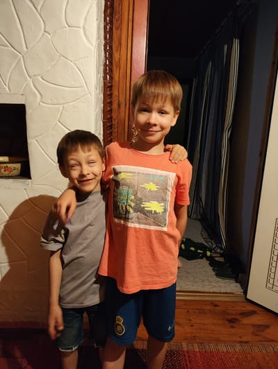 Two young boys, possibly brothers, stand together indoors. The boy on the left, wearing a gray t-shirt and denim shorts, has his eyes closed and a wide grin. The boy on the right, in an orange t-shirt and blue shorts, has his arm around the other boy's shoulder. They are positioned in front of a white, textured wall that appears to be part of a fireplace or stove. To the right of the boys is a wooden doorway with vertical blinds in the background. The flooring consists of wooden planks and a patterned rug. The lighting is dim, creating shadows on the wall. Based on the context of the provided image, the location is identified as the city of Khryashchevka, Russia.