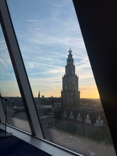 The image shows a view of the Martini Tower in Groningen, Netherlands, through a window. The tower is the focal point, towering over other buildings and appearing silhouetted against a sky with diffused sunlight. The window frame partially obscures the view, creating a framed perspective. The environment is urban, consisting of other buildings and structures. The sky transitions in color, suggesting either sunrise or sunset. There are no people visible in the image.